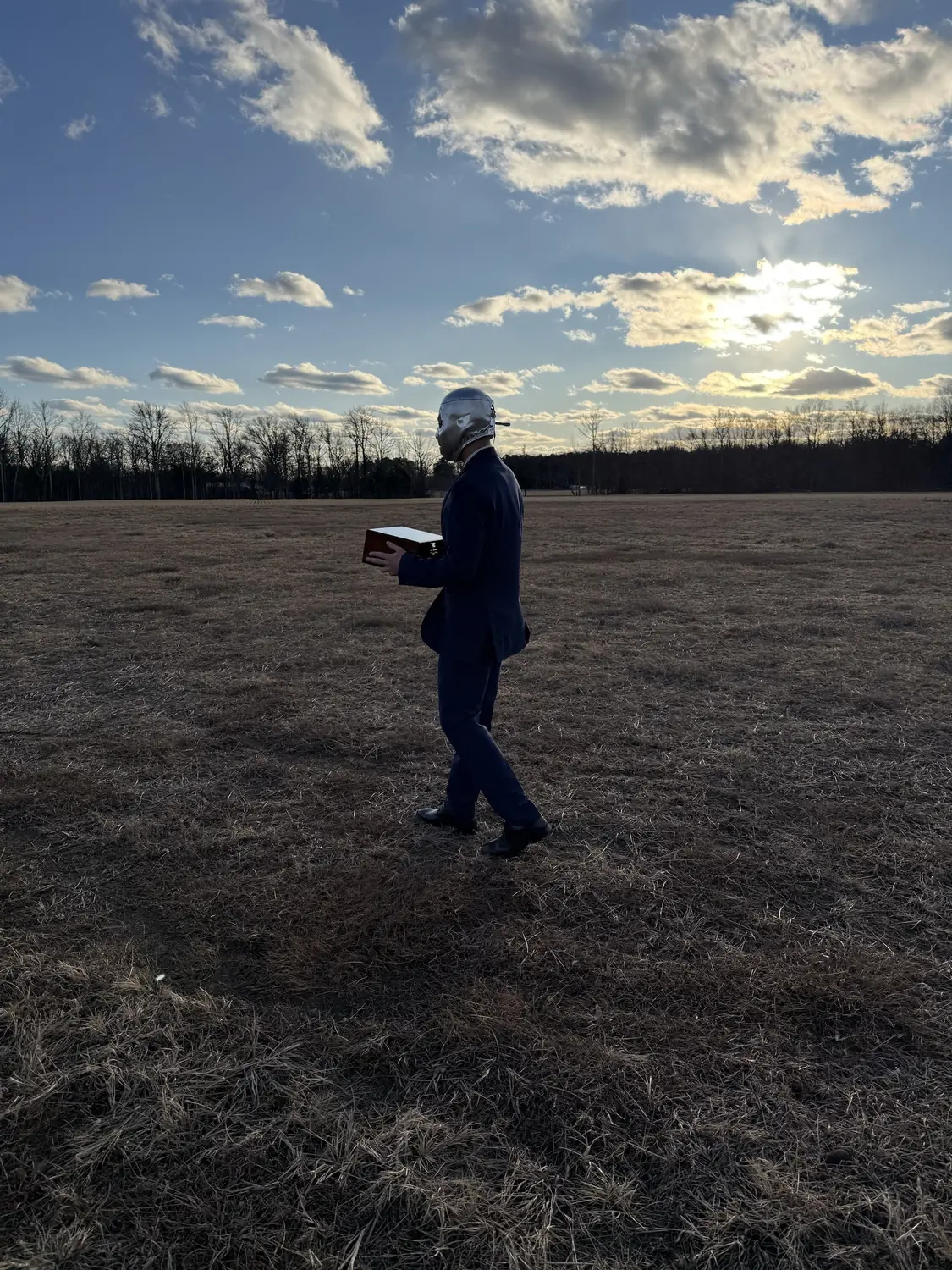 A figure in a dark suit and silver luchador mask carrying a wooden box across a field at golden hour