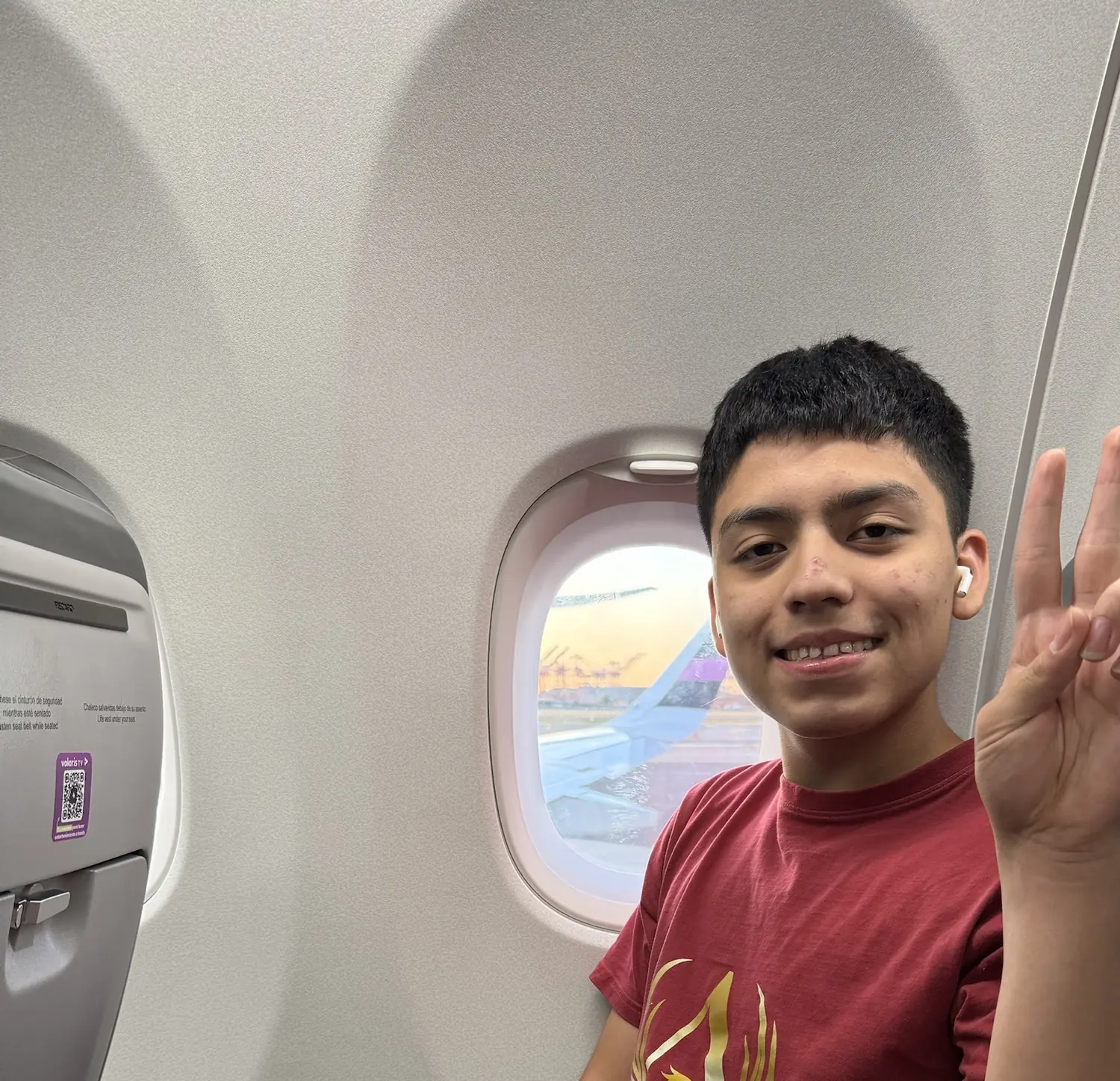 Adriel in a window seat on a plane at sunrise, wearing an Aureveris T-shirt and making a peace sign
